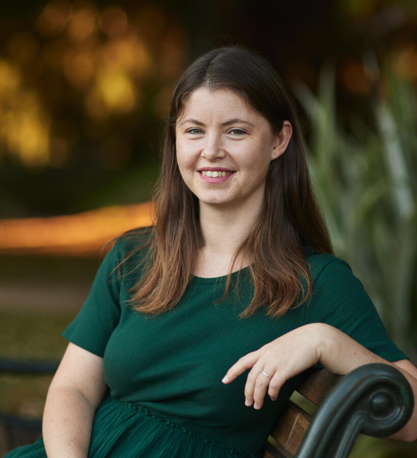Untitled Photo of Hannah Turrell. She has shoulder length straight brown hair, light skin, is wearing green and sitting on a park bench smiling.